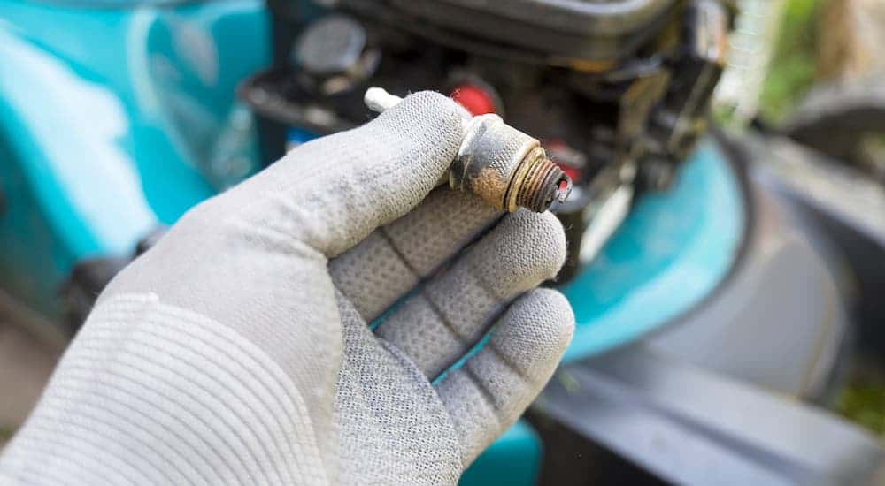 A gloved hand is holding a spark plug over a car's engine bay.