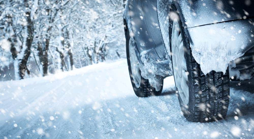 A closeup shows the wheels of a car driving on a snowy road.