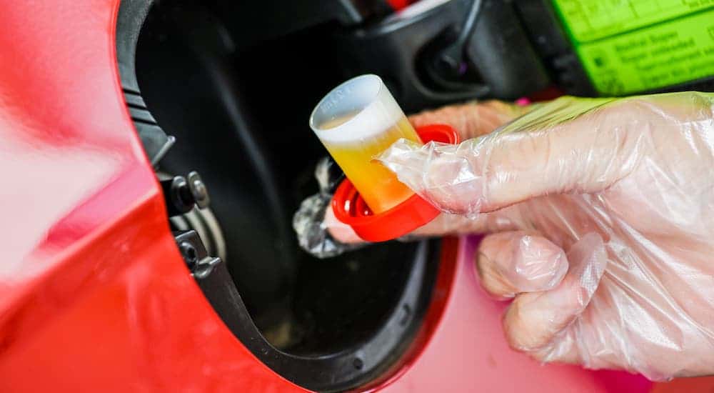 A closeup shows a gloved hand pouring an additive into the fuel filler neck of a red car.