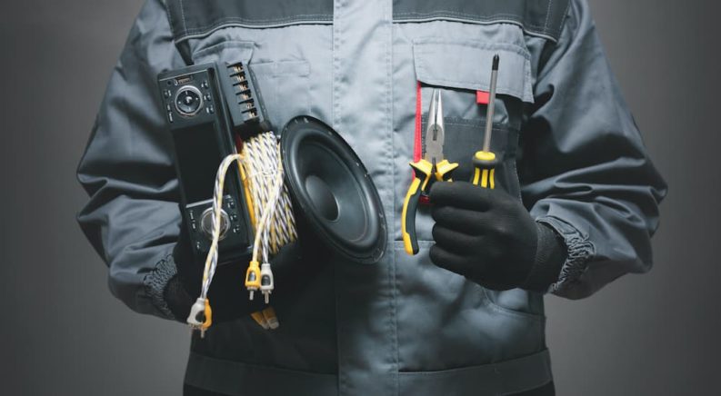 A close up shows a mechanic with his hands full of tools and a car speaker.