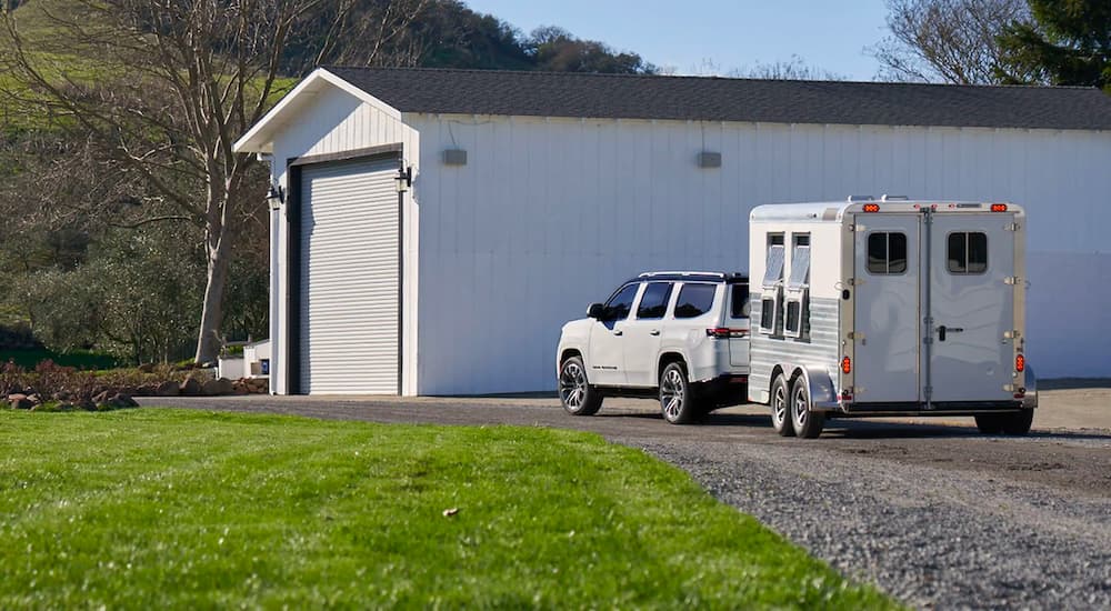A white 2022 Grand Wagoneer is shown towing a horse trailer past a white garage.
