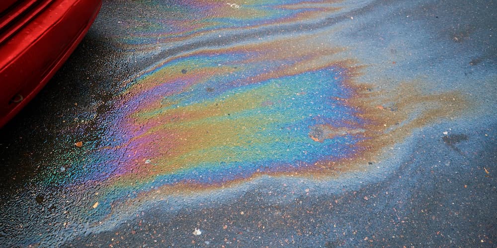 A rainbow colored gasoline leak is shown on pavement underneath a red car.