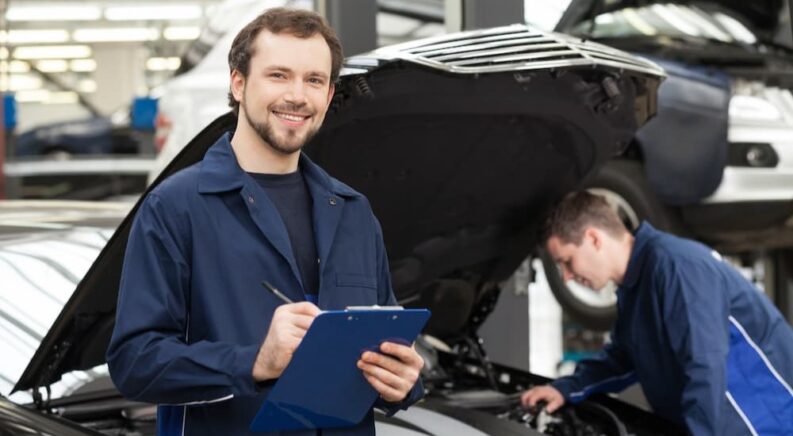 A mechanic is shown at an auto repair shop with a clipboard in his hands.