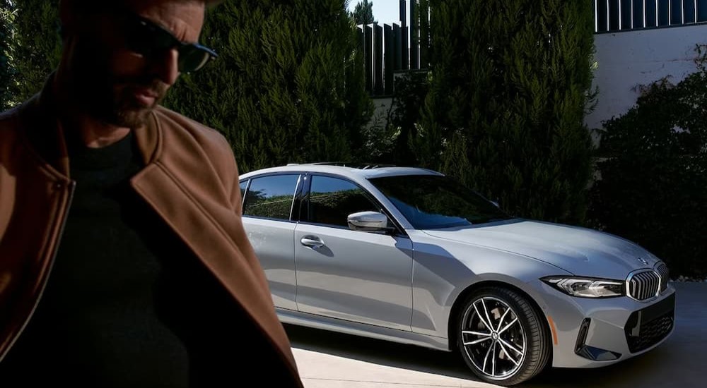 A silver 2024 BMW 3 Series is shown parked near a man wearing sunglasses.