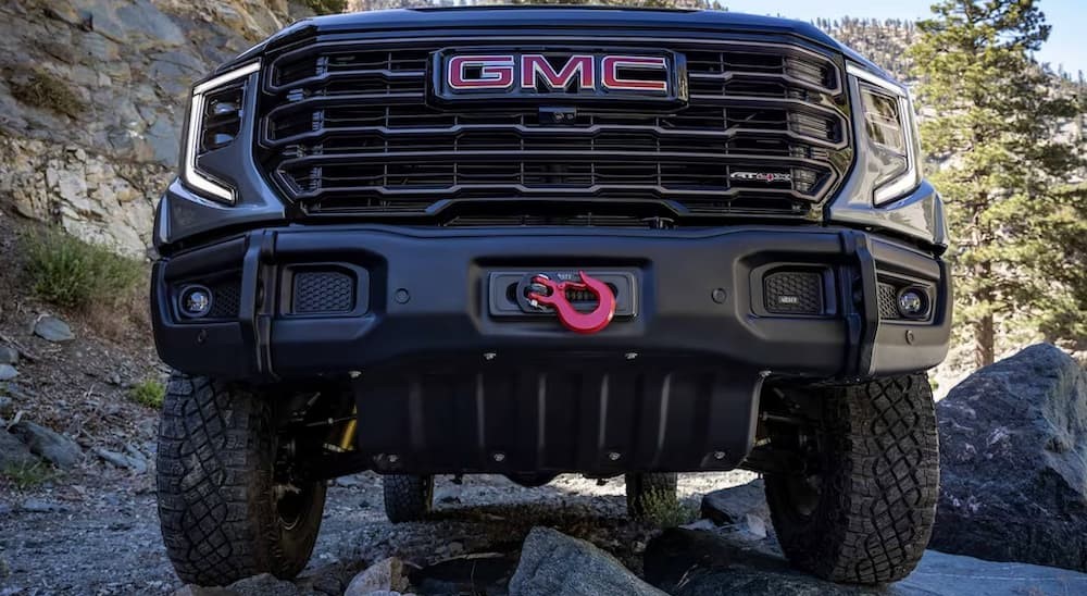One of many popular lifted trucks for sale, a close-up of the grille on a gray 2024 GMC Sierra 1500 AT4X, is shown parked off-road.