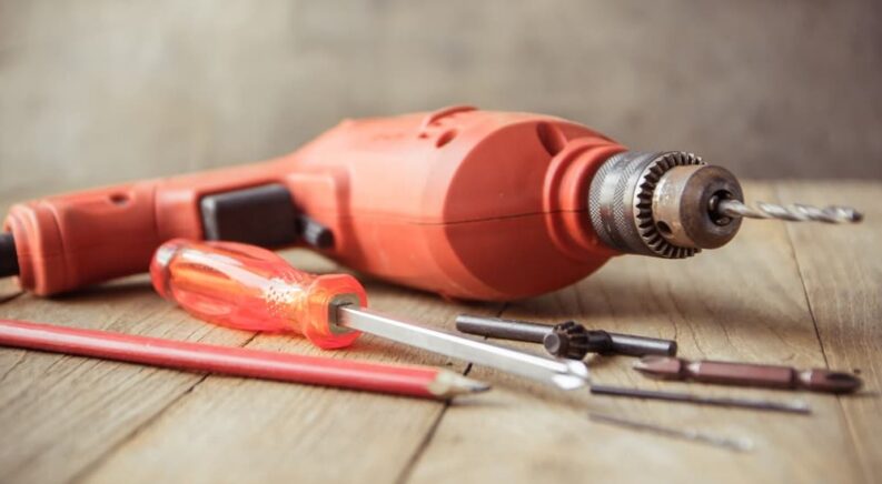 A red drill, bits, a screwdriver and a pencil are shown on a table.