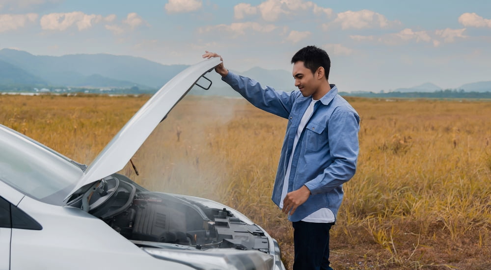 A man looking under the hood of an overheated white car.