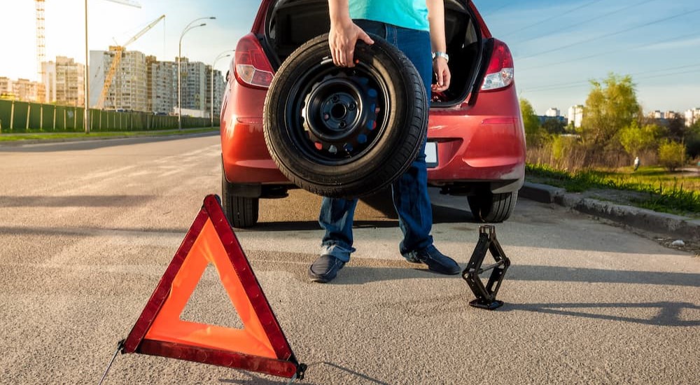 A person is holding a spare tire by a red car.